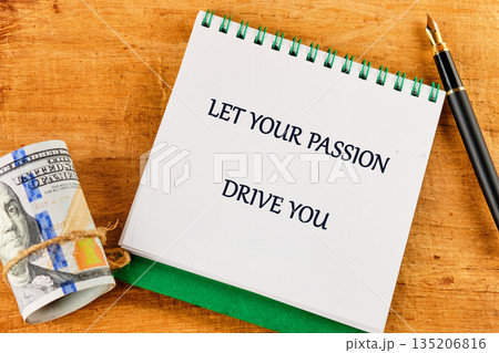 A roll of money and a sophisticated pen rest on a wooden desk beside a notepad showcasing motivational text, LET YOUR PASSION DRIVE 135206816