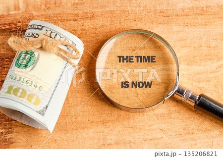 A roll of United States currency tied with twine rests beside a magnifying glass highlighting the message THE TIME IS NOW against a wood background 135206821