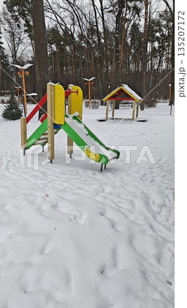 Snowy playground with colorful slide and play structure covered by fresh snow in winter park surrounded by trees. Concept of childhood, season change, silence, cold weather and abandoned recreation 135207172