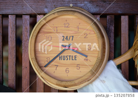 A wooden clock hangs on a wooden background. The clock face shows the numbers and the word Hustle. Its a unique piece of decor reminding us that time is of the essence 135207858