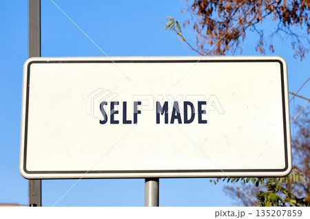 The scene shows a rectangular white sign with dark lettering that proclaims SELF MADE. It is attached to a metal post with bare tree branches and a vast blue sky in the background 135207859