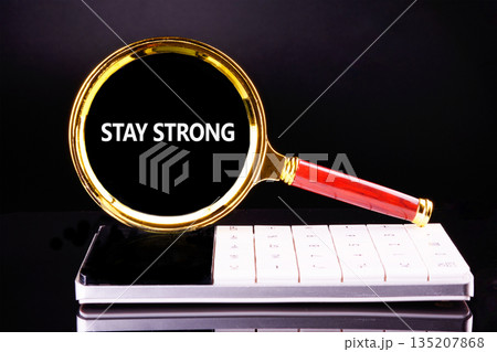 A gleaming gold and red magnifying glass highlights the powerful phrase STAY STRONG, resting artfully on a sleek, white calculator against a reflective, dark surface 135207868
