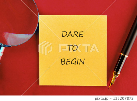 A bright yellow sticky note featuring the words DARE TO BEGIN rests on a crimson surface alongside a magnifying glass and a sleek fountain pen, beckoning a new start A bright yellow sticky note featuring the words DARE TO BEGIN rests on a crimson surface alongside a magnifying glass and a sleek fountain pen, beckoning a new start 135207872