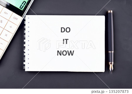 A notebook displays DO IT NOW in bold lettering. Placed on a dark surface with a calculator and a pen, urging immediate action and emphasizing the present 135207873