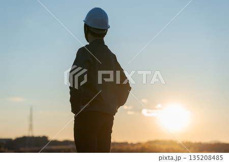 夕焼けをバックに立つ作業着の男性 夕焼けをバックに立つ作業着の男性 135208485