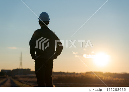 夕焼けをバックに立つ作業着の男性 135208486