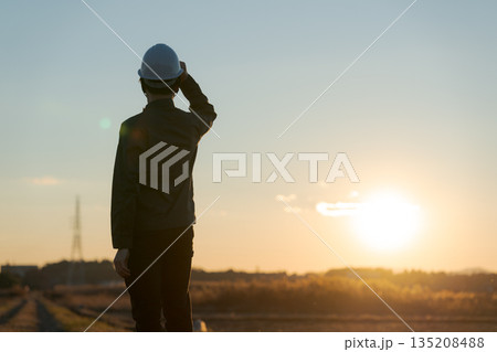夕焼けをバックに立つ作業着の男性 135208488