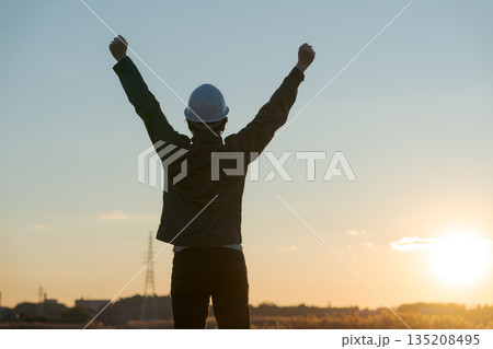 夕焼けをバックに万歳する作業着の男性 135208495