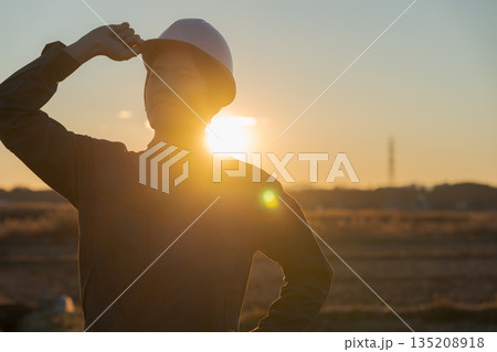 夕焼けをバックに立つ作業着の男性 135208918