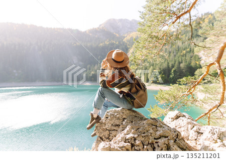 Back view young woman with backpack looking the view of the mountain lake in sunny weather. 135211201