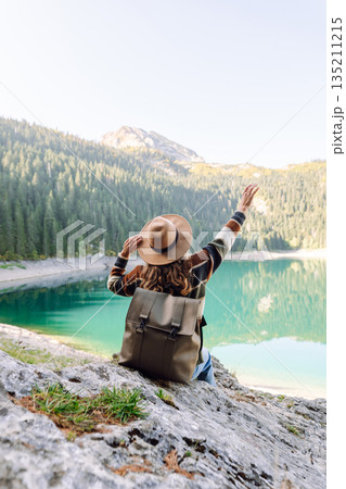 Back view young woman with backpack looking the view of the mountain lake in sunny weather. 135211215