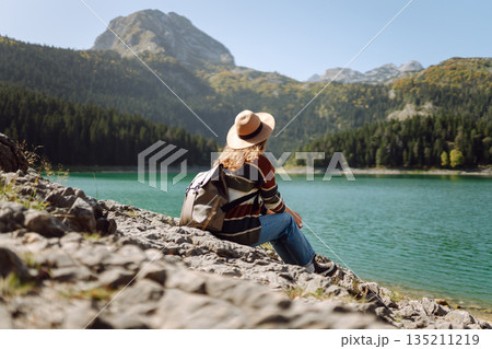Back view young woman with backpack looking the view of the mountain lake in sunny weather. Back view young woman with backpack looking the view of the mountain lake in sunny weather. 135211219