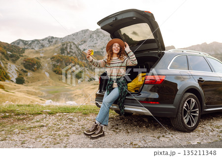 Young traveler woman taking a selfie by her car in the mountains during sunset. Lifestyle, travel 135211348