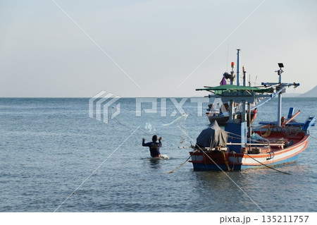 A man is fishing with a net in the sea, next to a colorful wooden fishing boat. A man is fishing with a net in the sea, next to a colorful wooden fishing boat. 135211757