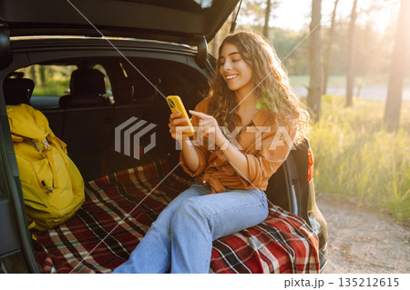 Young curly woman in stylish clothes in the trunk of a car in the forest and uses the phone. Young curly woman in stylish clothes in the trunk of a car in the forest and uses the phone. 135212615