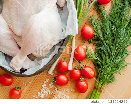 Whole raw chicken laying in the pan with parchment on the wooden table preparing for baking, surrounded by fresh vegetables. Whole raw chicken laying in the pan with parchment on the wooden table preparing for baking, surrounded by fresh vegetables. 135214251