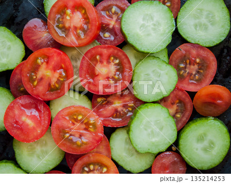 Bunch of sliced fresh red cherry tomatoes and cucumbers on the black background, top view veggies covered with water drops. Bunch of sliced fresh red cherry tomatoes and cucumbers on the black background, top view veggies covered with water drops. 135214253