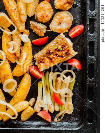 Oven pan with sliced baked potatoes, fish steaks and vegetables with onion on table. Oven pan with sliced baked potatoes, fish steaks and vegetables with onion on table. 135214261
