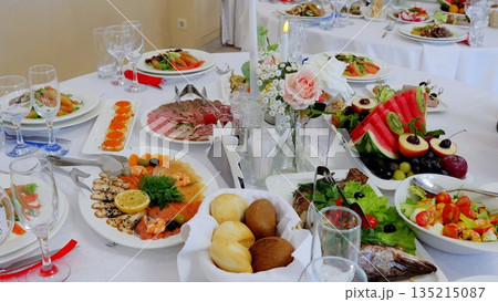 Beautifully decorated table is set for an event in a restaurant with various dishes of meat, fish, salads, fruits, vegetables, bread and flowers 135215087
