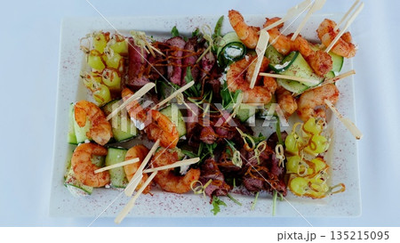 Miniature catering skewers presenting succulent shrimp, tender roast beef, cucumber, grapes, garnished with arugula and spices on pristine rectangular serving plate 135215095