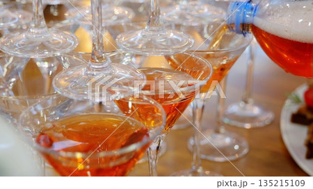 Professional bartender pouring bright orange cocktail into stacked glasses, creating festive celebration scene with dynamic drink presentation at bar or event venue Professional bartender pouring bright orange cocktail into stacked glasses, creating festive celebration scene with dynamic drink presentation at bar or event venue 135215109