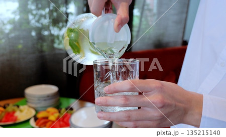 Waiter in white shirt pouring lemonade from lidded pitcher into crystal glass, fruity plates enhancing refreshing beverage presentation Waiter in white shirt pouring lemonade from lidded pitcher into crystal glass, fruity plates enhancing refreshing beverage presentation 135215140