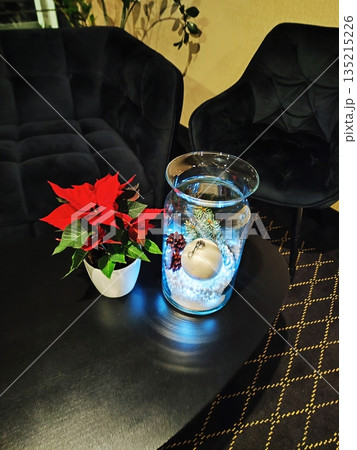 A black table with a poinsettia plant Euphorbia pulcherrima in a white pot sits next to a glass vase. The vase contains a white ornament, pine cones, and evergreen sprigs 135215226