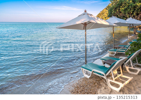 Beach chairs and umbrellas lined up on a serene tropical beach with crystal clear blue water Beach chairs and umbrellas lined up on a serene tropical beach with crystal clear blue water 135215313
