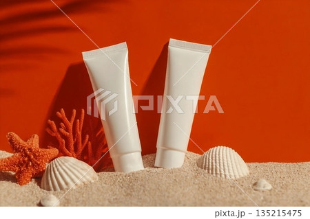 Two white cosmetic tubes are arranged creatively with seashells, coral, and starfish on sand against an orange backdrop, evoking a beachy vibe. Two white cosmetic tubes are arranged creatively with seashells, coral, and starfish on sand against an orange backdrop, evoking a beachy vibe. 135215475