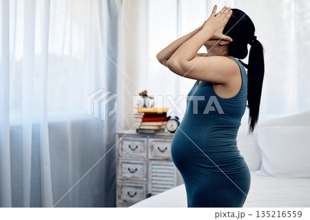 Pregnant woman in blue dress standing indoors with hands covering face feeling stress and fatigue in bedroom 135216559