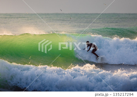 Surfer man rides on wave in cold winter ocean with evening sunlight Surfer man rides on wave in cold winter ocean with evening sunlight 135217294