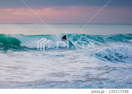 Surfer in wetsuit rides the waves in ocean with sunset tones Surfer in wetsuit rides the waves in ocean with sunset tones 135217298