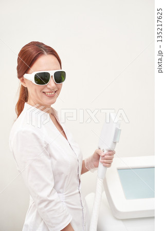 Female caucasian adult in white coat holding cosmetic laser equipment, smiling 135217625