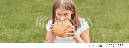 Caucasian young girl enjoying large bread loaf outdoors in green park setting 135217824