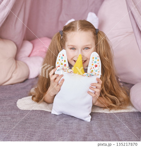Happy caucasian female child with pigtails holding unicorn pillow in cozy pink 135217870