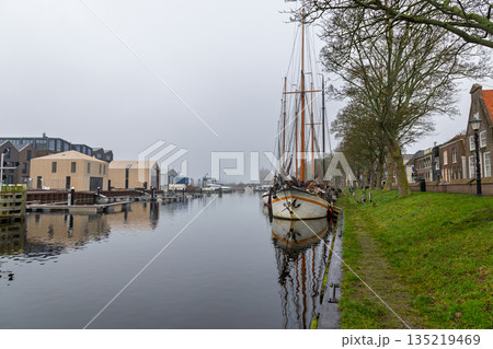 Misty Dutch canal scene leafless trees, docked sailboat, traditional homes and modern buildings reflect in calm water, blending heritage and contemporary waterfront design. 135219469