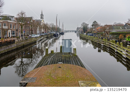 Misty Dutch canal sailboats moored beside brick homes, church steeple, and quiet streets with bikes and cars reflect in calm water, blending heritage and serenity. Misty Dutch canal sailboats moored beside brick homes, church steeple, and quiet streets with bikes and cars reflect in calm water, blending heritage and serenity. 135219470