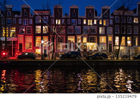 Amsterdam canal at night gabled buildings with glowing windows, parked cars, and string lit boat reflect warmly on tranquil water. Amsterdam canal at night gabled buildings with glowing windows, parked cars, and string lit boat reflect warmly on tranquil water. 135219479