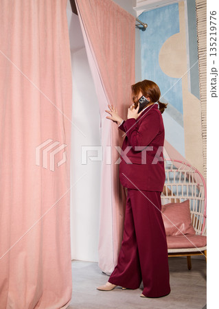 Woman in maroon outfit checks through curtain calmly during call Woman in maroon outfit checks through curtain calmly during call 135219776