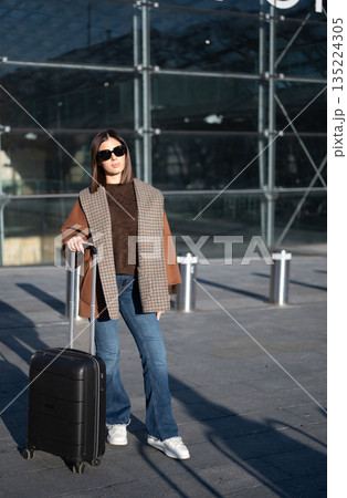 Woman standing outdoors near a glass facade, wearing sunglasses and warm stylish clothes, holding a black suitcase, ready for a journey or urban exploration 135224305