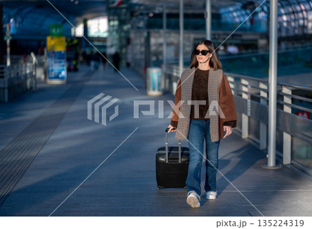 Woman wearing sunglasses and a brown jacket with plaid detail walking and pulling a black suitcase, arriving at a bright, modern airport or train station during travel Woman wearing sunglasses and a brown jacket with plaid detail walking and pulling a black suitcase, arriving at a bright, modern airport or train station during travel 135224319