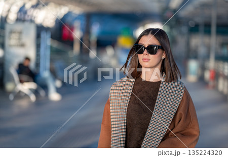 Chic brunette in sunglasses and a patterned lapel coat walks confidently down an urban street, embodying modern, elegant city style and casual confidence in daylight Chic brunette in sunglasses and a patterned lapel coat walks confidently down an urban street, embodying modern, elegant city style and casual confidence in daylight 135224320