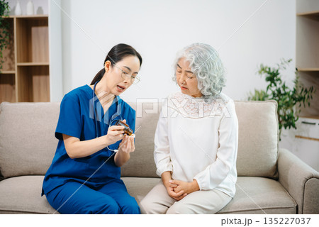Young nurse holding a bottle of pills and discussing treatment with an elderly Asian woman during a medical consultation Young nurse holding a bottle of pills and discussing treatment with an elderly Asian woman during a medical consultation 135227037