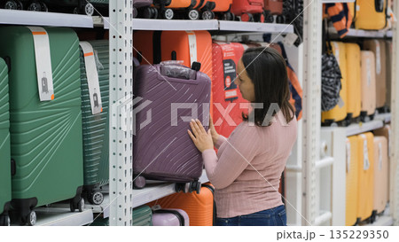 Young woman checking a purple suitcase on a store shelf, inspecting surface material and handle for quality, travel purchase and vacation concept Young woman checking a purple suitcase on a store shelf, inspecting surface material and handle for quality, travel purchase and vacation concept 135229350