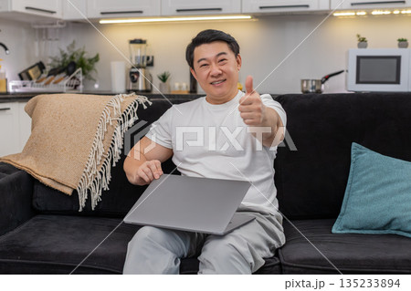 Asian man closes laptop after tasks exhales stretches smiles pride shows big thumbs up toward camera Asian man closes laptop after tasks exhales stretches smiles pride shows big thumbs up toward camera 135233894