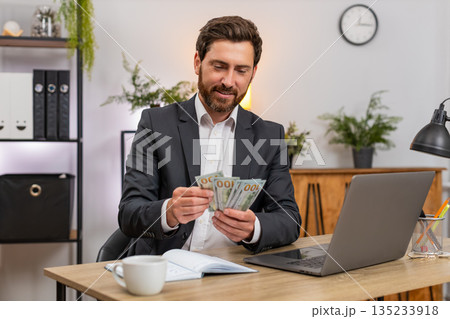 Middle-aged businessman counts cash dollars records budget on laptop writes notes in notebook plans Middle-aged businessman counts cash dollars records budget on laptop writes notes in notebook plans 135233918