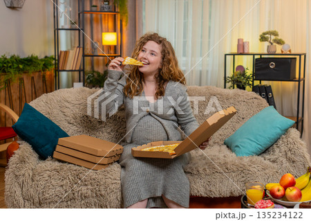 Pregnant woman opening pizza box at home, tasting slice with satisfied smile, enjoying comfort food 135234122