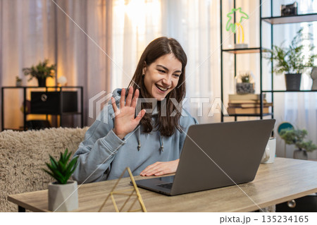 Happy young woman sitting at table talking to friend by video call using laptop wave hand at home Happy young woman sitting at table talking to friend by video call using laptop wave hand at home 135234165