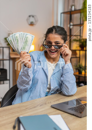 Young woman freelancer putting on party glasses holding fan of cash sitting at home office table 135234183