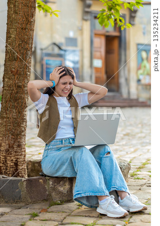 Caucasian young woman using laptop browsing loses lottery results, bad news sitting in city street Caucasian young woman using laptop browsing loses lottery results, bad news sitting in city street 135234211